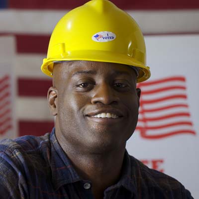 A smiling man wearing a construction hard hat with an American flag behind him
